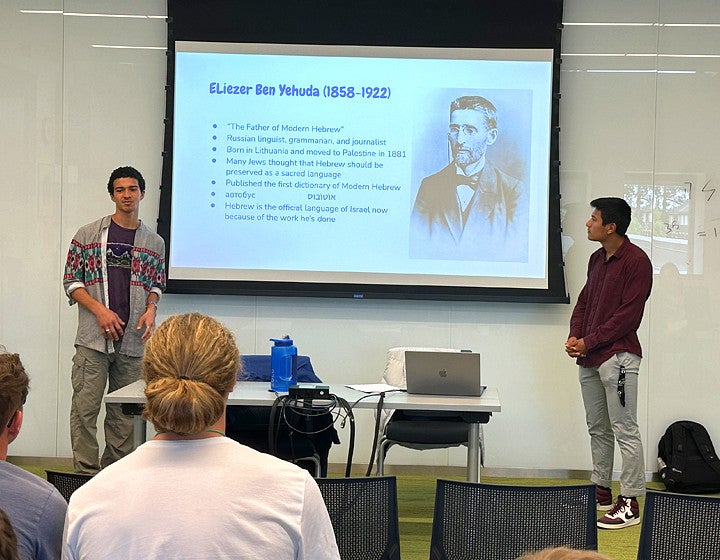 Two students are giving a presentation on stage with a screen behind them