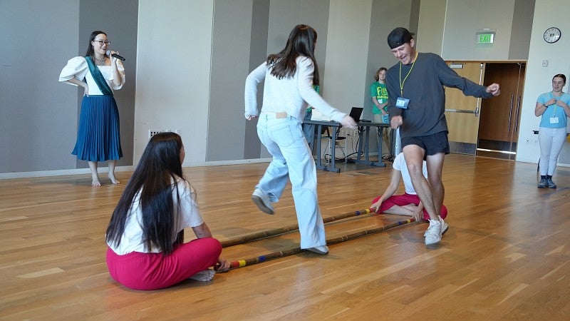 students jumping over two long sticks on the ground while two other students hold the sticks