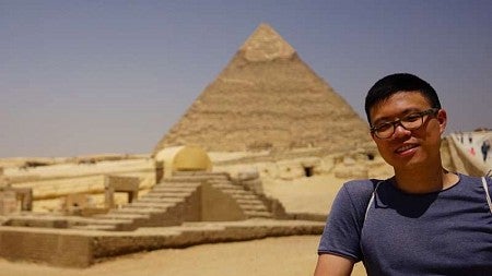 man with glasses standing to the right of a great pyramid in the background