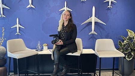 woman sitting on a stool holding a camera with a wall of model airplanes behind her