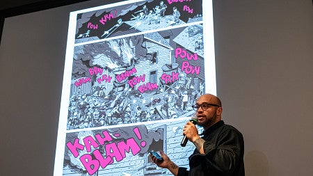 man with a microphone standing in front of a screen featuring a comics page
