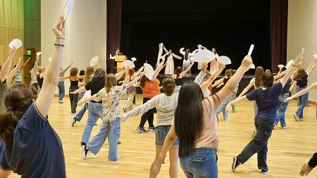 students stand in a gymnasium facing a stage holding up fans and dancing during a traditional Chinese fan dance
