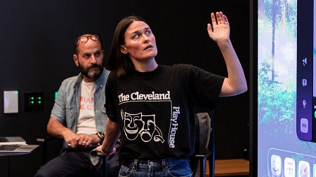 woman standing in front of a lecture hall gesturing toward the screen behind her