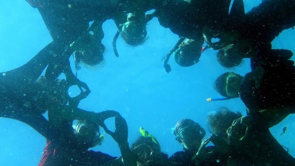 Students underwater in diving gear linking arms in circle while throwing the O