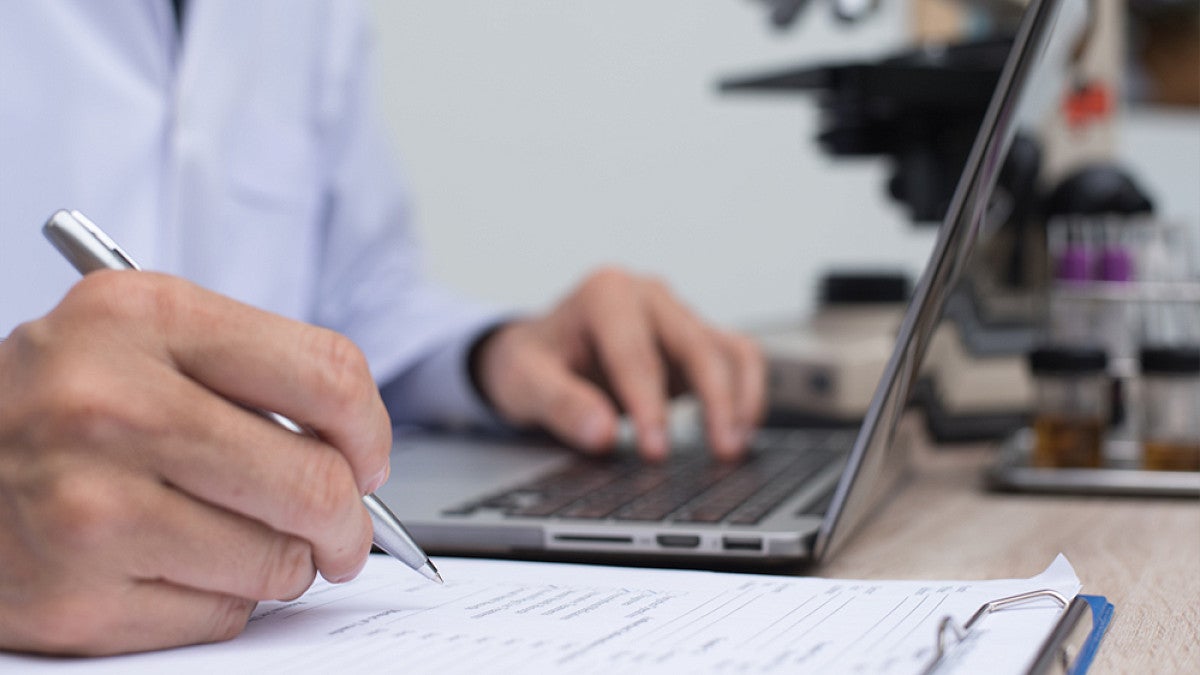 A person writing on a note pad while using a laptop sitting at a table