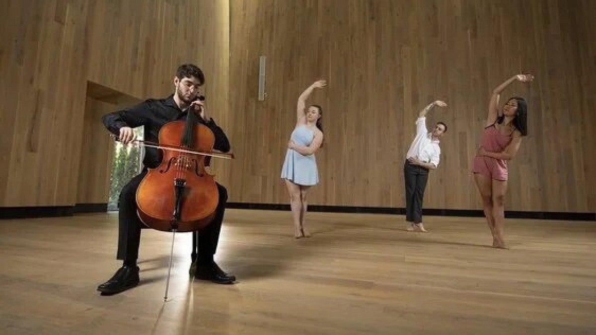 Person playing a cello with 3 other people dancing ballet in the background