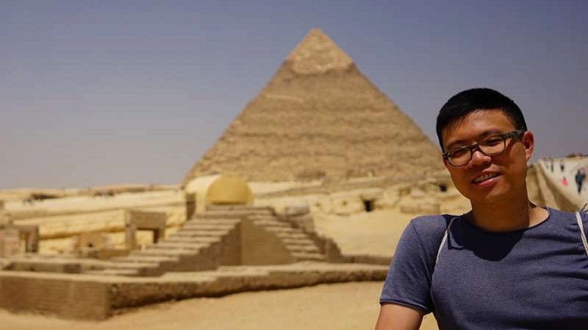 man with glasses standing to the right of a great pyramid in the background