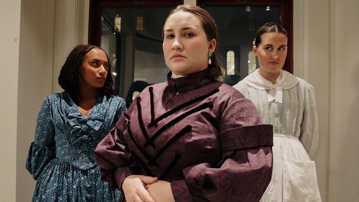 three women wearing 19th century dresses stand in slightly different positions with suspicious expressions