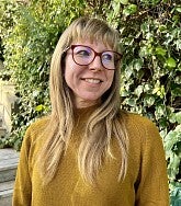 photo of woman with long hair and glasses standing in front of greenery