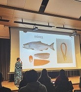 woman holding a microphone standing on a stage in front of a screen with images of fish and beaded necklace